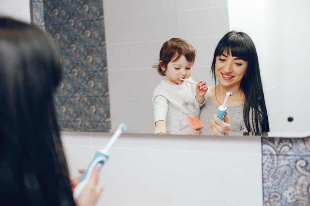 A little kid brushing his teeth along with her mother 